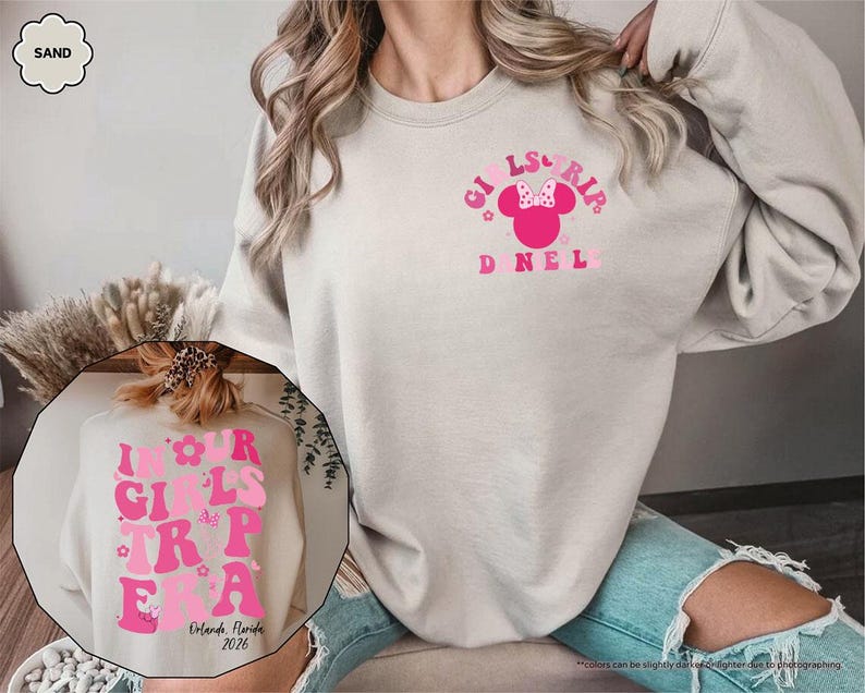 Peut inclure: Un sweat-shirt couleur sable avec l'inscription "GIRLS TRIP" en rose, orn&eacute; d'un motif de t&ecirc;te de Minnie Mouse. Le sweat-shirt porte &eacute;galement l'inscription "IN OUR GIRLS TRIP ERA" et "Orlando, Florida 2026" dans une image circulaire.