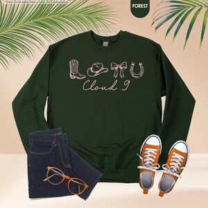 May include: A dark green sweatshirt featuring a pink illustration of cowboy-themed items and the words "Cloud 9". The image also shows a pair of blue jeans, orange trainers, and glasses. The sweatshirt is the focal point.
