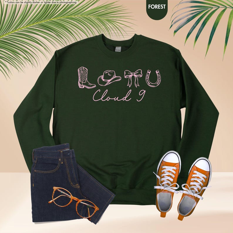 May include: A dark green sweatshirt featuring a pink illustration of cowboy-themed items and the words "Cloud 9". The image also shows a pair of blue jeans, orange trainers, and glasses. The sweatshirt is the focal point.