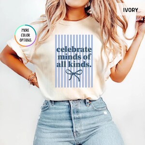 Puede incluir: Camiseta de color marfil con el texto azul "celebrate minds of all kinds" sobre un fondo a rayas azul claro. Un lazo azul est&aacute; debajo del texto. La camiseta tiene mangas cortas.