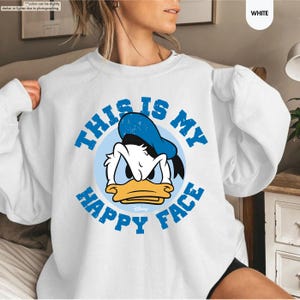 Op de afbeelding: Witte sweatshirt met een afbeelding van Donald Duck en de tekst "THIS IS MY HAPPY FACE" in blauw. De cartoon eend draagt een blauwe hoed en heeft een boze uitdrukking. De sweatshirt is een casual stijl.