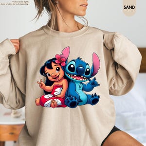 Puede incluir: Sudadera color arena con un gr&aacute;fico de Lilo y Stitch. Lilo se representa con cabello negro, un vestido rojo y blanco y una flor rosa. Stitch es azul con orejas grandes. La sudadera tiene mangas largas.