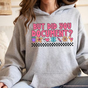 Puede incluir: Una sudadera gris claro con el texto "BUT DID YOU DOCUMENT?" en rosa y blanco. El dise&ntilde;o incluye gr&aacute;ficos de tem&aacute;tica m&eacute;dica como una taza de caf&eacute;, una venda, un estetoscopio, un coraz&oacute;n y una jeringa. La sudadera tiene un bolsillo delantero y una capucha con cord&oacute;n.