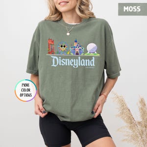 Op de afbeelding: Een mosgroen t-shirt met een Disneyland Resort-afbeelding. De afbeelding toont een reuzenrad, kasteel en Spaceship Earth. Het woord "Disneyland" is geschreven in een gestileerd lettertype.