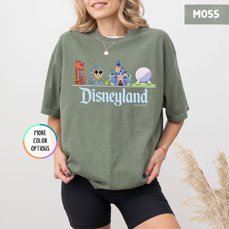 Op de afbeelding: Een mosgroen t-shirt met een Disneyland Resort-afbeelding. De afbeelding toont een reuzenrad, kasteel en Spaceship Earth. Het woord "Disneyland" is geschreven in een gestileerd lettertype.