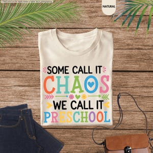 May include: Cream-colored t-shirt with the text "SOME CALL IT CHAOS WE CALL IT PRESCHOOL" in colorful letters. The shirt is folded on a wooden surface next to a pair of blue jeans and a brown purse.