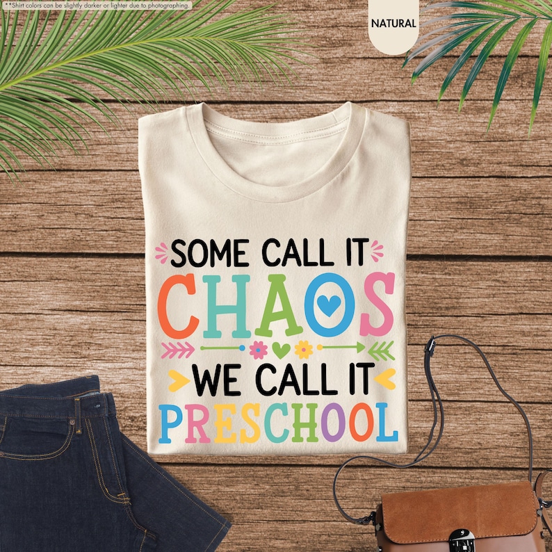 May include: Cream-colored t-shirt with the text "SOME CALL IT CHAOS WE CALL IT PRESCHOOL" in colorful letters. The shirt is folded on a wooden surface next to a pair of blue jeans and a brown purse.