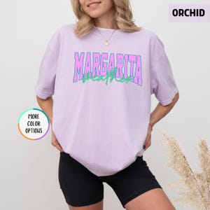 May include: A light purple t-shirt with the words "MARGARITA WEATHER" in pink and green lettering. The shirt is paired with black shorts. The image also includes a "MORE COLOR OPTIONS" button and the word "ORCHID".