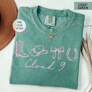 May include: Light green Comfort Colors t-shirt with a pink graphic of a cowboy boot, hat, bow, and horseshoe. The text "Cloud 9" is written in pink script below the graphics. A gold necklace is draped across the shirt.