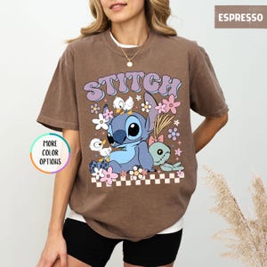 May include: Espresso-colored t-shirt featuring a cartoon graphic of Stitch with the word "STITCH" in a retro font. The design includes flowers and ducks. The shirt has a crew neck and short sleeves. "MORE COLOR OPTIONS" is printed on a circular button.