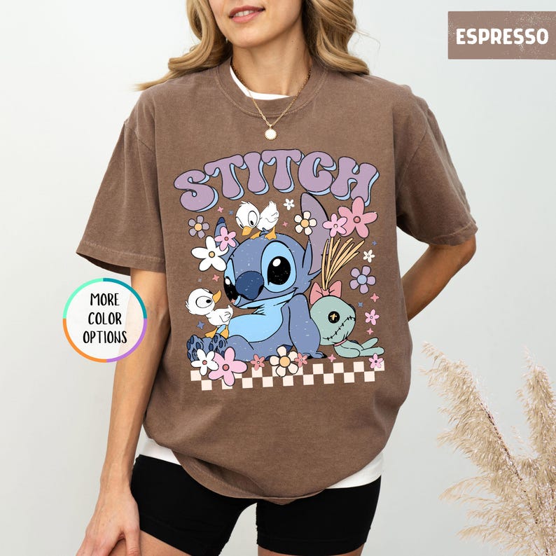 May include: Espresso-colored t-shirt featuring a cartoon graphic of Stitch with the word "STITCH" in a retro font. The design includes flowers and ducks. The shirt has a crew neck and short sleeves. "MORE COLOR OPTIONS" is printed on a circular button.