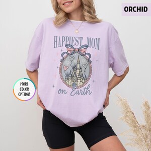 May include: Orchid-colored t-shirt with the text "Happiest Mom on Earth" above a castle graphic. The graphic includes a pink bow, Mickey Mouse balloons, and a castle. The shirt has short sleeves and a round neckline.