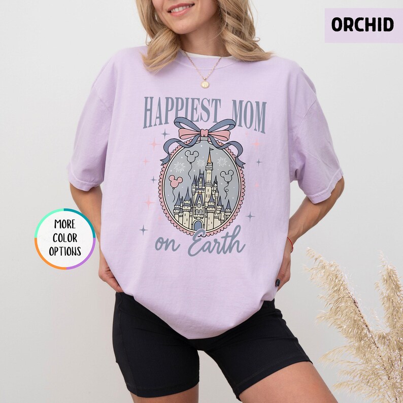 May include: Orchid-colored t-shirt with the text "Happiest Mom on Earth" above a castle graphic. The graphic includes a pink bow, Mickey Mouse balloons, and a castle. The shirt has short sleeves and a round neckline.