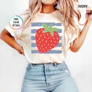 May include: An ivory t-shirt featuring a large red strawberry graphic with a green stem centered over horizontal blue distressed stripes. The shirt includes a circular graphic with the text MORE COLOR OPTIONS and a label in the corner reading IVORY.