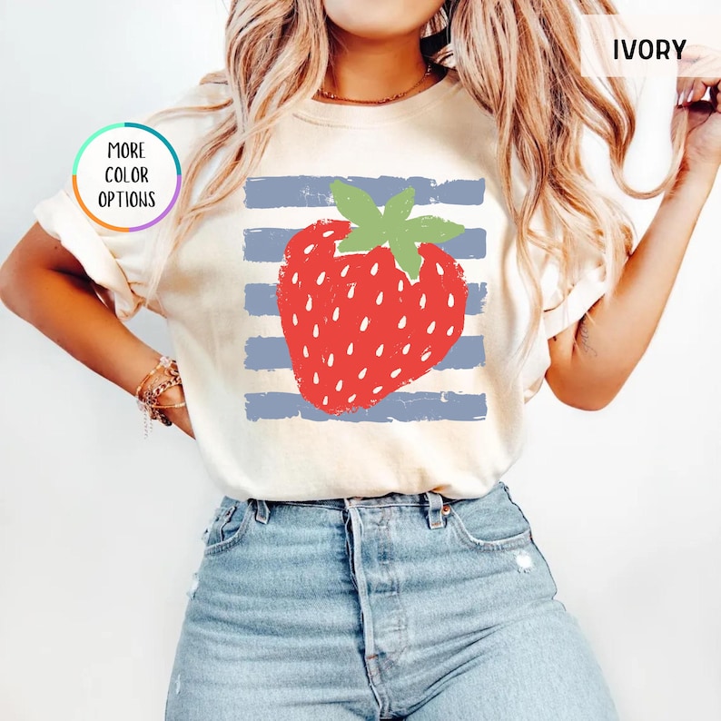 May include: An ivory t-shirt featuring a large red strawberry graphic with a green stem centered over horizontal blue distressed stripes. The shirt includes a circular graphic with the text MORE COLOR OPTIONS and a label in the corner reading IVORY.
