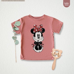 May include: A heather mauve t-shirt featuring a Minnie Mouse graphic. The graphic shows Minnie Mouse in a seated pose with her hands under her chin. She is wearing a red bow with white polka dots. A wooden baby rattle is next to the shirt.