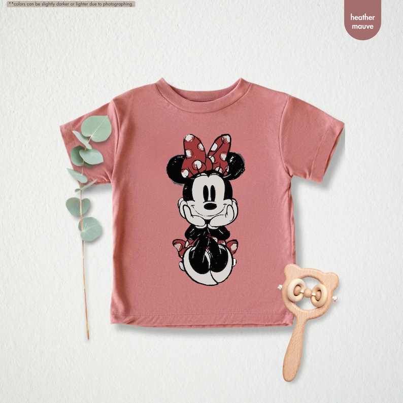 May include: A heather mauve t-shirt featuring a Minnie Mouse graphic. The graphic shows Minnie Mouse in a seated pose with her hands under her chin. She is wearing a red bow with white polka dots. A wooden baby rattle is next to the shirt.