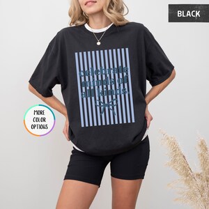 Puede incluir: Camiseta negra con la frase "celebrate minds of all kinds" en azul claro, sobre un fondo de rayas azules verticales. El dise&ntilde;o incluye un lazo en la parte inferior. La camiseta tiene cuello redondo y mangas cortas.
