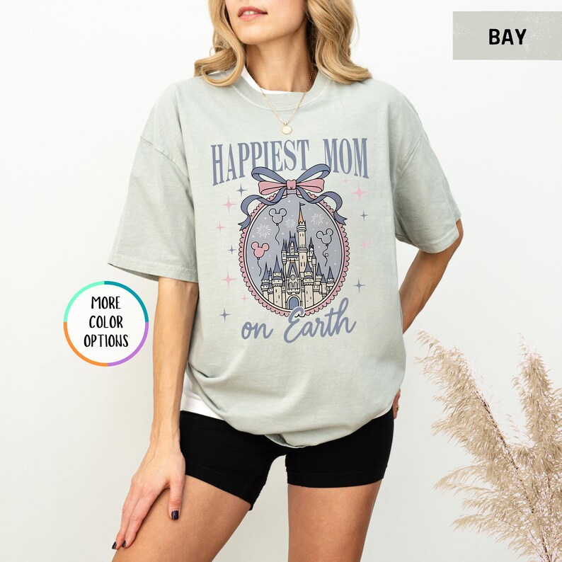 May include: A light green t-shirt with the text "Happiest Mom on Earth" and a castle graphic. The graphic includes a pink bow, balloons, and a castle. The shirt is a relaxed fit and has short sleeves.