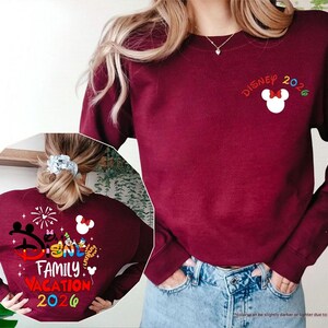 Puede incluir: Una sudadera de cuello redondo color granate con un peque&ntilde;o icono de cabeza de Mickey Mouse en blanco con un lazo rojo y el texto Disney 2026 en el pecho. La parte trasera muestra un gr&aacute;fico grande con personajes de Disney, fuegos artificiales y el texto Disney Family Vacation 2026.