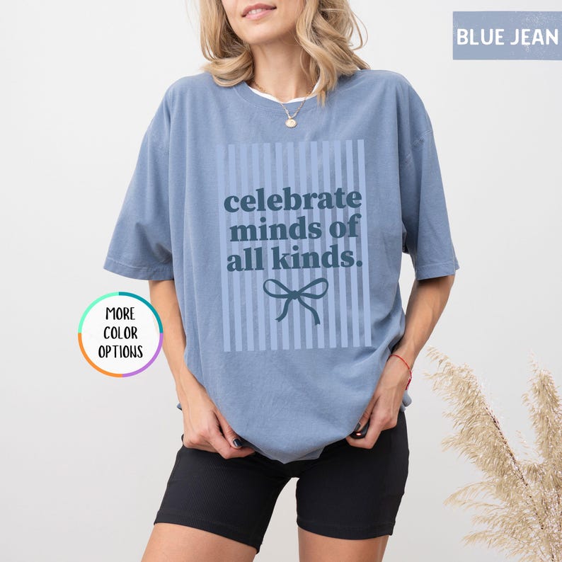 Puede incluir: Una camiseta azul vaquero con el texto "celebrate minds of all kinds" en un dise&ntilde;o cuadrado. El dise&ntilde;o incluye rayas verticales y un lazo. La camiseta es de color liso y tiene mangas cortas.