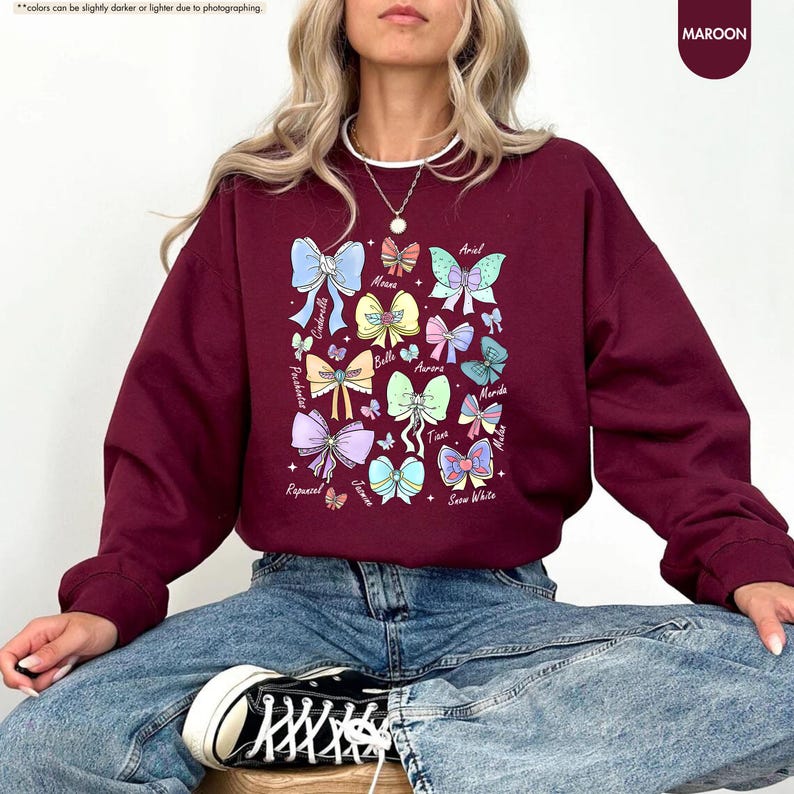 May include: Maroon sweatshirt with a colourful bow design featuring character names such as Cinderella and Ariel. The bows are in various colours, including blue, pink, and yellow. The sweatshirt has long sleeves and a crew neck.