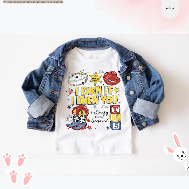 Peut inclure: Un t-shirt blanc superpos&eacute; sous une veste en jean bleu. Le t-shirt pr&eacute;sente un motif avec le texte I KNEW IT I KNEW YOU infinity and beyond, accompagn&eacute; d'illustrations d'une &eacute;toile de sh&eacute;rif, d'un chapeau rouge, de bottes et de cubes.