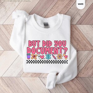 Puede incluir: Sudadera blanca con la frase "BUT DID YOU DOCUMENT?" en letras rosas y naranjas. El dise&ntilde;o incluye gr&aacute;ficos m&eacute;dicos como una venda, un estetoscopio y una jeringa, con un borde a cuadros blanco y negro.
