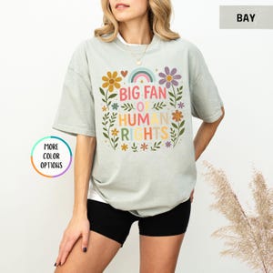 May include: A light green t-shirt with the text "BIG FAN OF HUMAN RIGHTS" in colorful letters, surrounded by floral and rainbow graphics. The shirt is paired with black shorts.