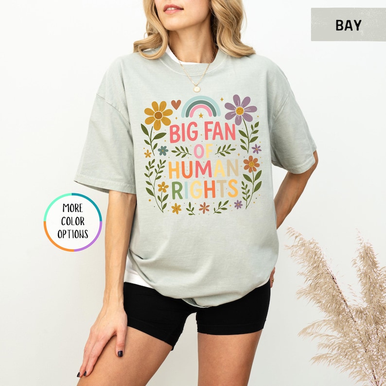 May include: A light green t-shirt with the text "BIG FAN OF HUMAN RIGHTS" in colorful letters, surrounded by floral and rainbow graphics. The shirt is paired with black shorts.