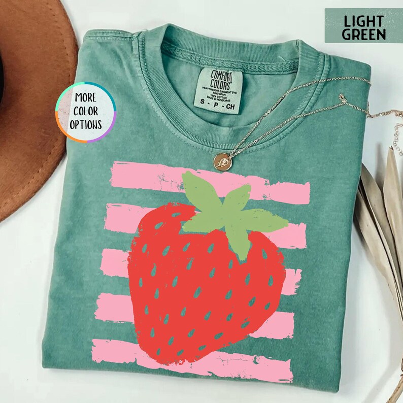 May include: A light green short-sleeve t-shirt featuring a large red strawberry graphic centered over horizontal pink brushstroke stripes. The shirt has a crew neckline with a Comfort Colors brand label and a thin gold-tone chain necklace.