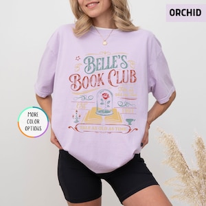 Pu&ograve; includere: T-shirt color orchidea con la scritta "Belle's Book Club" in un design vintage. La grafica include una rosa in una cupola di vetro, un libro aperto e la frase "Tale as Old as Time". La maglietta ha maniche corte.
