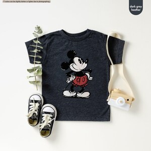 May include: Dark grey heather t-shirt featuring a classic Mickey Mouse graphic. The cartoon character is depicted in black, white, and red. The image also shows a pair of black and white sneakers, a small wooden camera, and a canvas bag.
