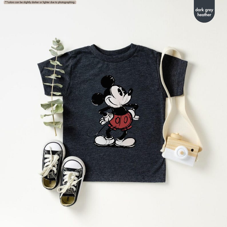 May include: Dark grey heather t-shirt featuring a classic Mickey Mouse graphic. The cartoon character is depicted in black, white, and red. The image also shows a pair of black and white sneakers, a small wooden camera, and a canvas bag.