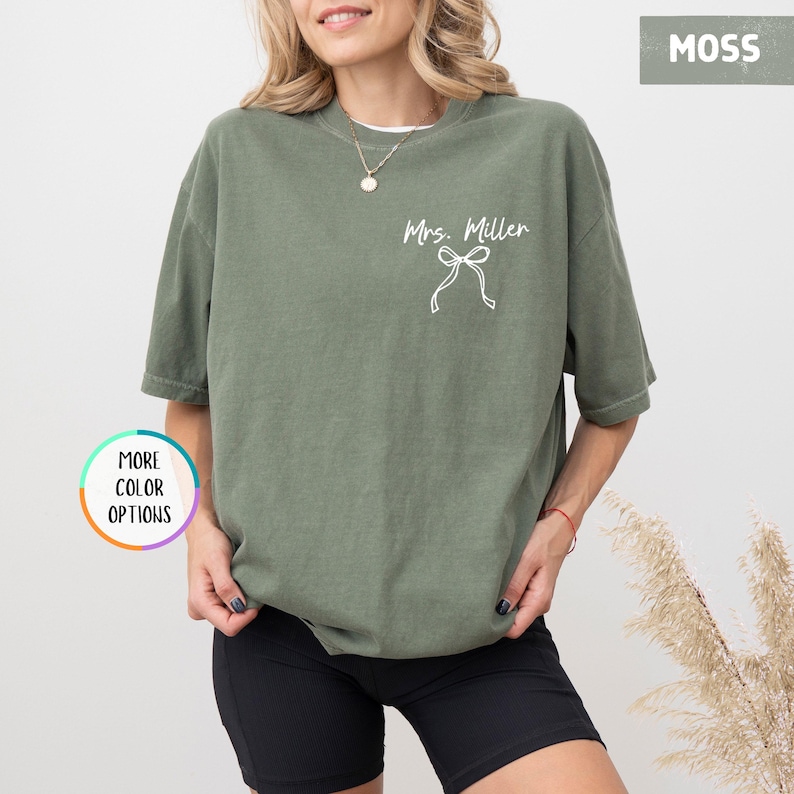 Puede incluir: Una camiseta verde musgo con el texto "Mrs. Miller" y un lazo blanco. La camiseta tiene mangas cortas y un corte holgado. Las palabras "More Color Options" est&aacute;n en un c&iacute;rculo en el lado izquierdo de la imagen.