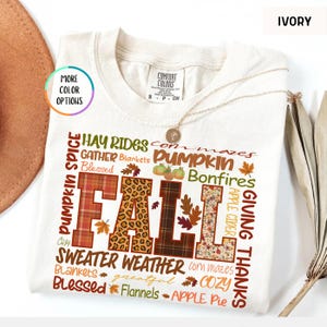 以下が含まれることがあります： 秋をテーマにした言葉と柄のコラージュで「FALL」の文字がデザインされた、アイボリー色のクルーネックTシャツ。チェック柄、ヒョウ柄、花柄、茶色の模様が特徴です。「Pumpkin Spice」、「Sweater Weather」、「Giving Thanks」の文字も表示されています。
