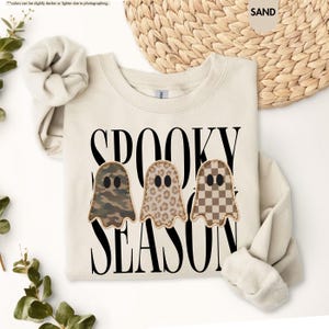 May include: A sand-colored crewneck sweatshirt featuring the black text SPOOKY SEASON and three ghost-shaped appliques. The ghosts have distinct patterns: one in camouflage, one in leopard print, and one in a brown and white checkerboard pattern.