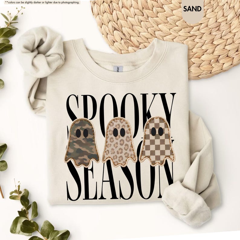 May include: A sand-colored crewneck sweatshirt featuring the black text SPOOKY SEASON and three ghost-shaped appliques. The ghosts have distinct patterns: one in camouflage, one in leopard print, and one in a brown and white checkerboard pattern.
