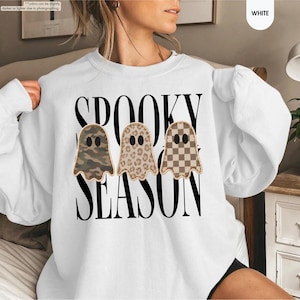 May include: A white crewneck sweatshirt featuring the black text SPOOKY SEASON. Three ghost illustrations are centered, each with a different pattern: one with green and brown camouflage, one with a tan leopard print, and one with a brown checkerboard.