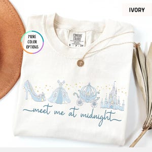 May include: Ivory-colored t-shirt with a light blue Cinderella-themed graphic. The design includes a glass slipper, ball gown, carriage, and castle, with the text "meet me at midnight" in a script font. A gold necklace is draped on the shirt.