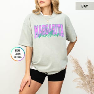 May include: A light green t-shirt with the words "MARGARITA WEATHER" in pink and green lettering. The shirt is a relaxed fit and has short sleeves. The model is wearing black shorts.