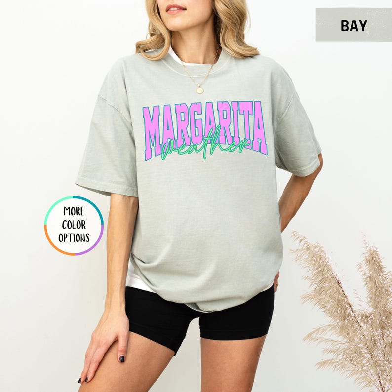 May include: A light green t-shirt with the words "MARGARITA WEATHER" in pink and green lettering. The shirt is a relaxed fit and has short sleeves. The model is wearing black shorts.