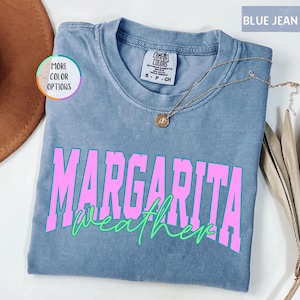 May include: Blue jean colored t-shirt with the words "MARGARITA weather" in pink and green. The shirt has a crew neck and short sleeves. A gold necklace with a pendant is draped across the shirt.