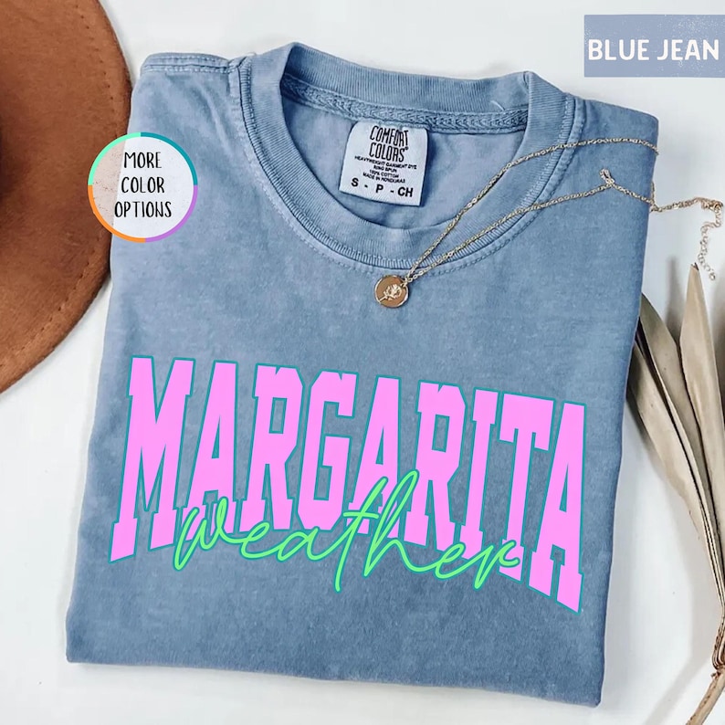 May include: Blue jean colored t-shirt with the words "MARGARITA weather" in pink and green. The shirt has a crew neck and short sleeves. A gold necklace with a pendant is draped across the shirt.