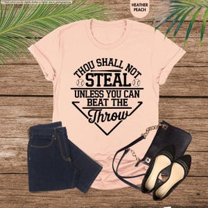 May include: Heather peach t-shirt with the text "Thou Shall Not Steal Unless You Can Beat The Throw" in black. The image also shows a pair of dark blue jeans, a black purse, and black flats.