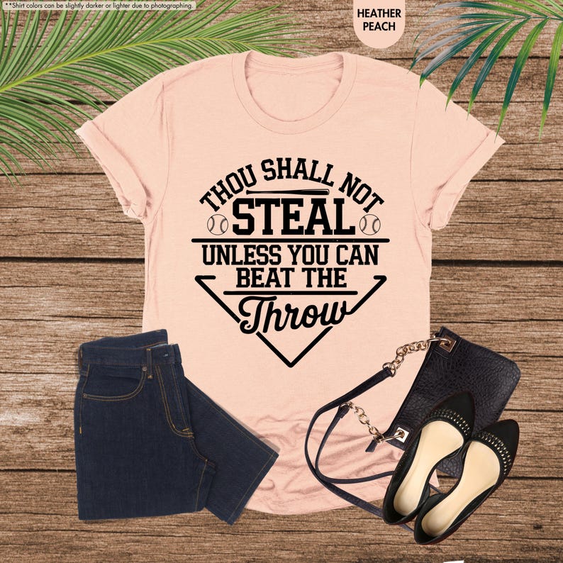 May include: Heather peach t-shirt with the text "Thou Shall Not Steal Unless You Can Beat The Throw" in black. The image also shows a pair of dark blue jeans, a black purse, and black flats.