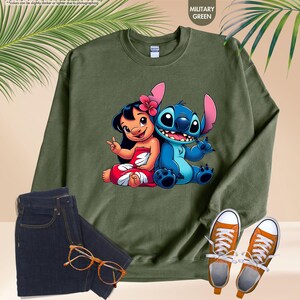 Puede incluir: Sudadera verde militar con un gr&aacute;fico de Lilo y Stitch. La parte superior de manga larga con cuello redondo se combina con vaqueros de lavado oscuro, zapatillas naranjas y gafas. Una hoja de palma est&aacute; en el fondo.