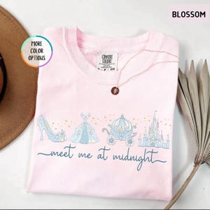 May include: A light pink Comfort Colors sweatshirt with a Cinderella theme. The design includes a blue glass slipper, ball gown, carriage, and castle, with the words "meet me at midnight" in script. A necklace with a pendant is draped on the shirt.