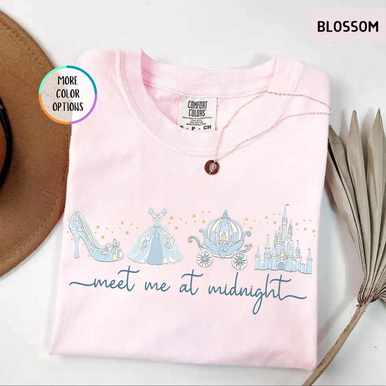 May include: A light pink Comfort Colors sweatshirt with a Cinderella theme. The design includes a blue glass slipper, ball gown, carriage, and castle, with the words "meet me at midnight" in script. A necklace with a pendant is draped on the shirt.