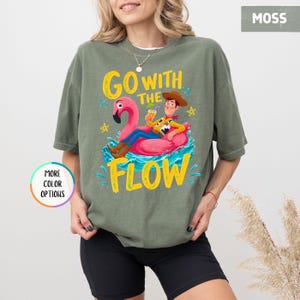 Puede incluir: Una camiseta de manga corta color verde musgo con un gr&aacute;fico de un vaquero sentado en un flotador de flamenco rosa sobre agua azul. El texto "GO WITH THE FLOW" aparece en letras amarillas, junto a peque&ntilde;as estrellas amarillas y salpicaduras.